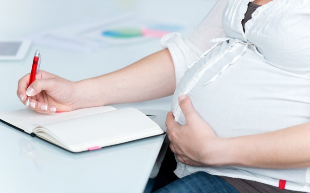 Pregnant Businesswoman Writing In Book At Desk
