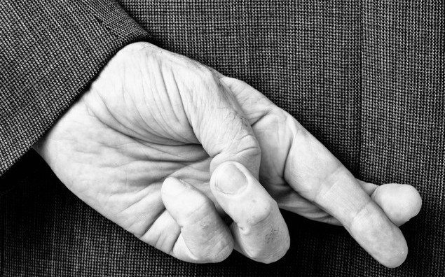 A B/W conceptial image of a business man with his fingers crossed behind his back.